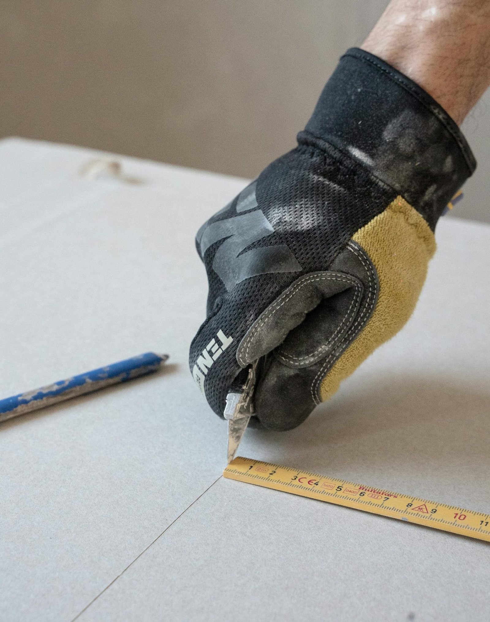 A gloved hand using a cutter and ruler to cut plasterboard for construction work.
